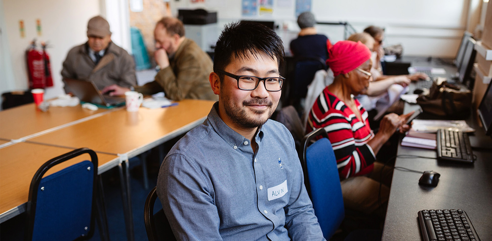 A person working in a Digital Inclusion Hub smiles at the camera. There's a digital skills session happening in the background with people using computers and tablets.